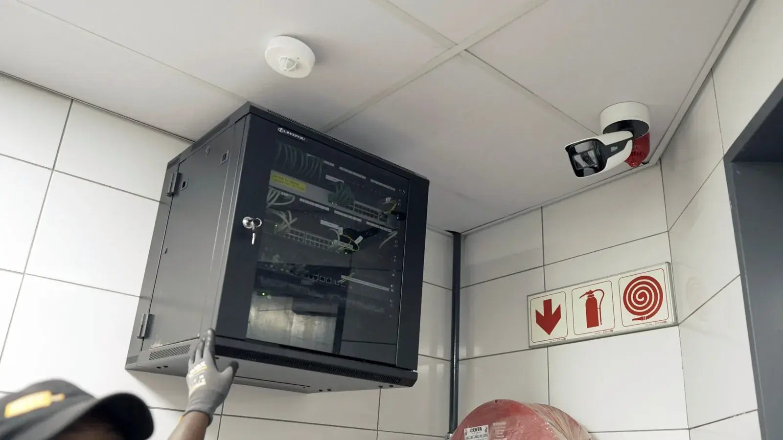 Ceiling smoke detector beside a CCTV bullet camera and network cabinet, with fire hose reel and extinguisher signs