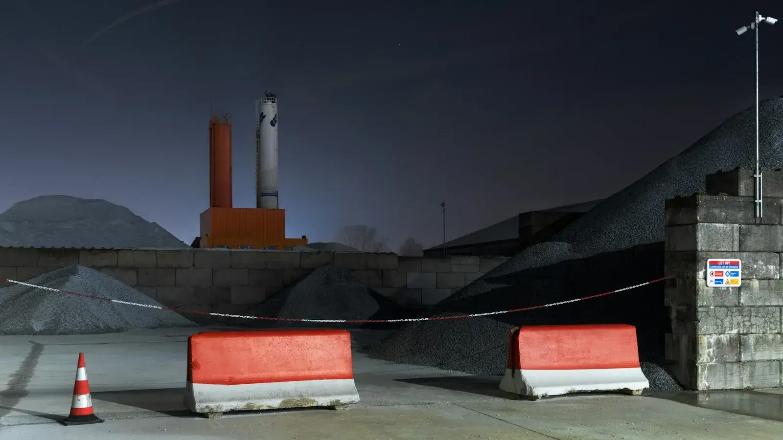 Industrial site at night with red-and-white concrete barriers, silos and a lighting mast